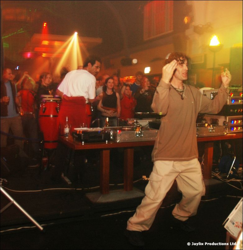 A person dances in front of a DJ table with conga drums, with a crowd and lights behind