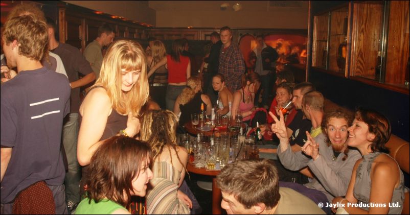 People seated and standing around tables with glasses in a busy bar, some pulling faces at the camera