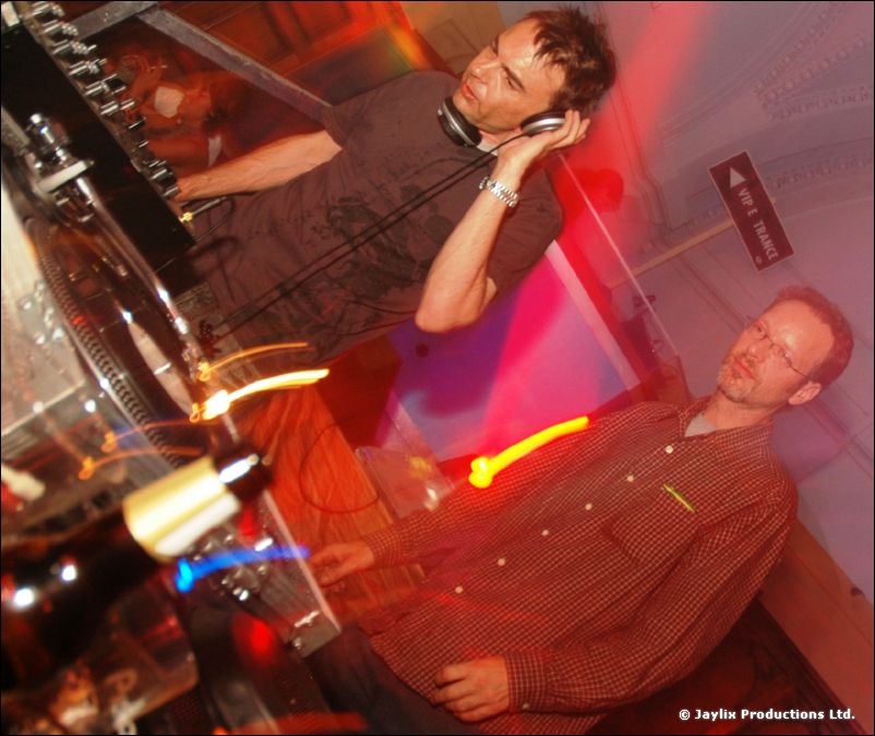 Two DJs with headphones at the decks in a tilted shot, with red and yellow light streaks