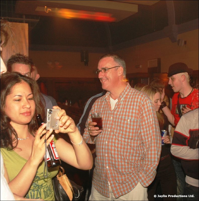 A man in a check shirt holding a drink and a woman photographing with a small camera in a crowded room