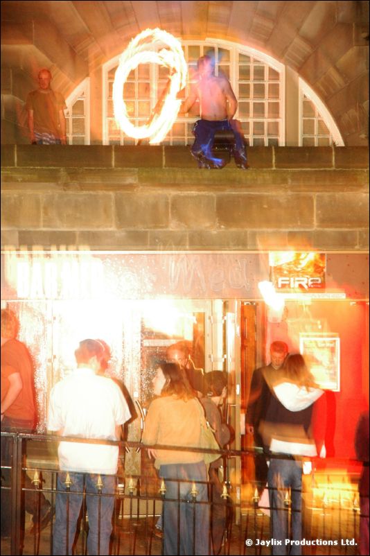A person on a stone ledge above an archway swings a glowing ring of fire, with people below