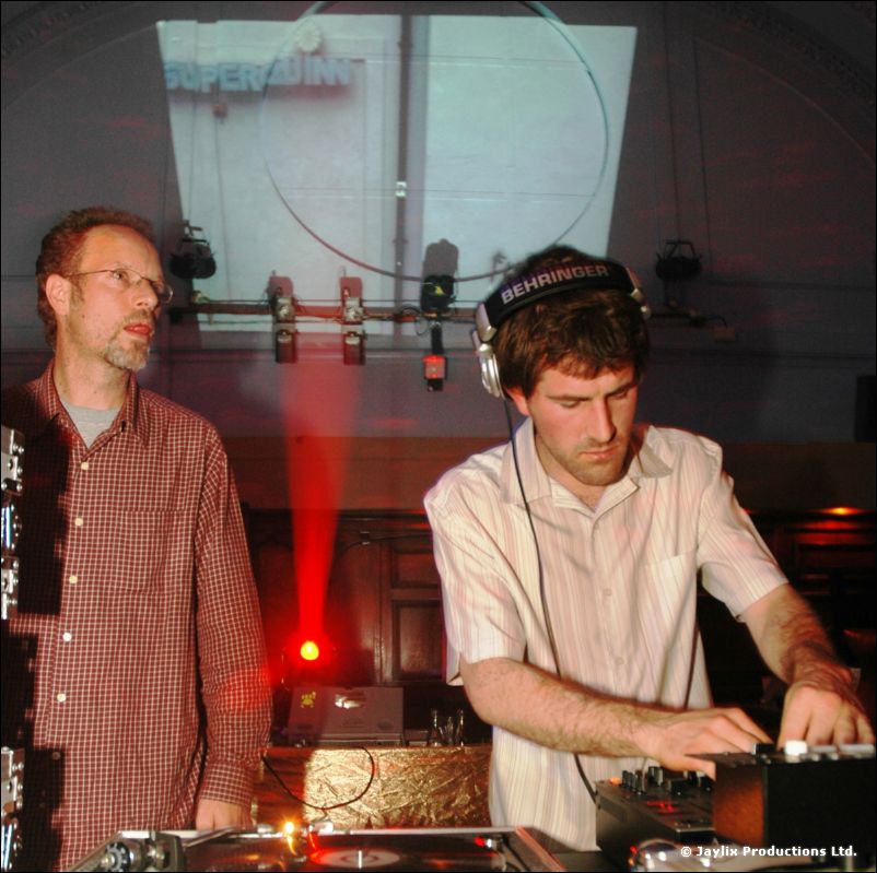 A DJ in a white shirt and Behringer headphones at the decks, with a man in a checked shirt beside him
