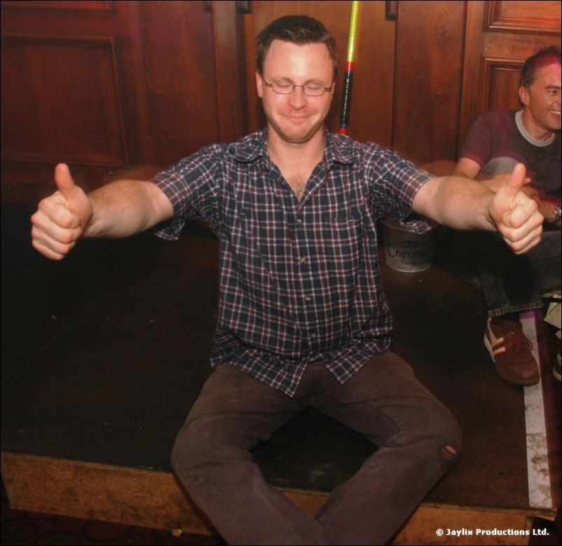 A man in a checked shirt with glasses gives two thumbs up, seated in a wood panelled room