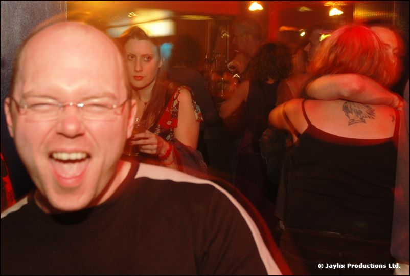 A bald man with glasses laughs at the camera, with people embracing behind him