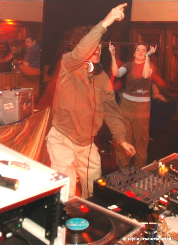 A DJ in headphones points upward beside the decks, with another person dancing behind