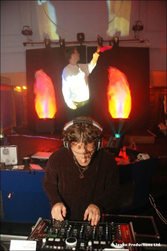 A DJ at a mixer in front of two glowing flame lights and a person in white with raised arms