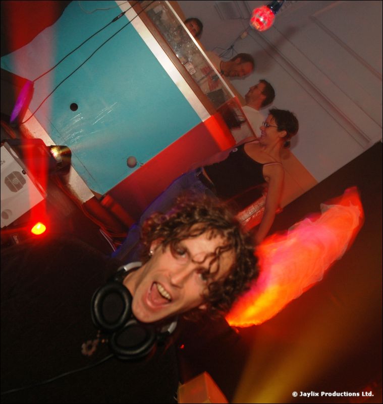 A tilted close-up of a DJ with headphones around his neck and curly hair, with a glowing red shape beside