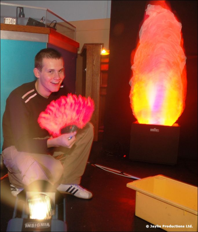 A smiling person holds a pink feather fan beside a glowing flame light