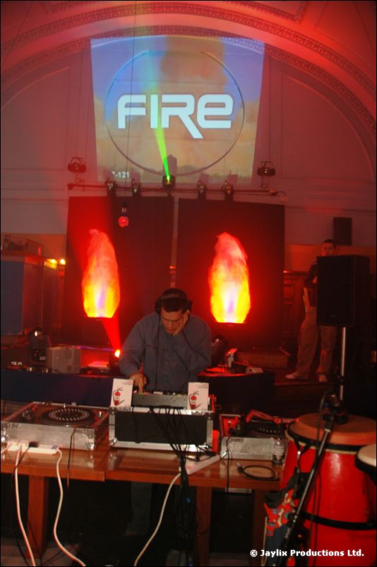 A DJ at turntables beside two glowing flame lights, with a projected "FIRE" logo and congas