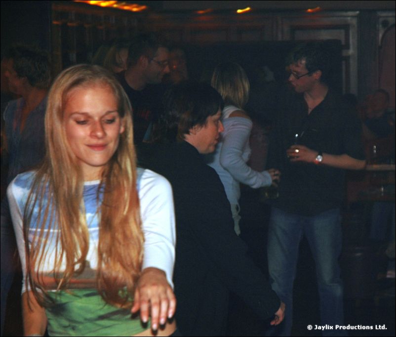 A woman with long hair in a white top looks down as people stand behind her in a dark bar