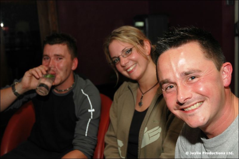 Two men and a woman with glasses sit close and smile, one man drinking from a bottle