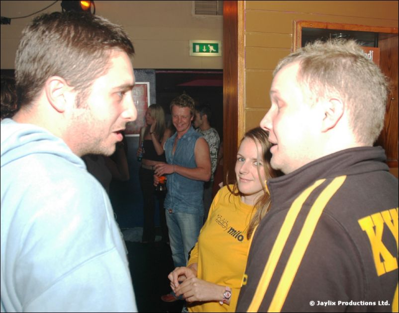 Two men in hooded and striped tops talk closely, with a woman in yellow behind them