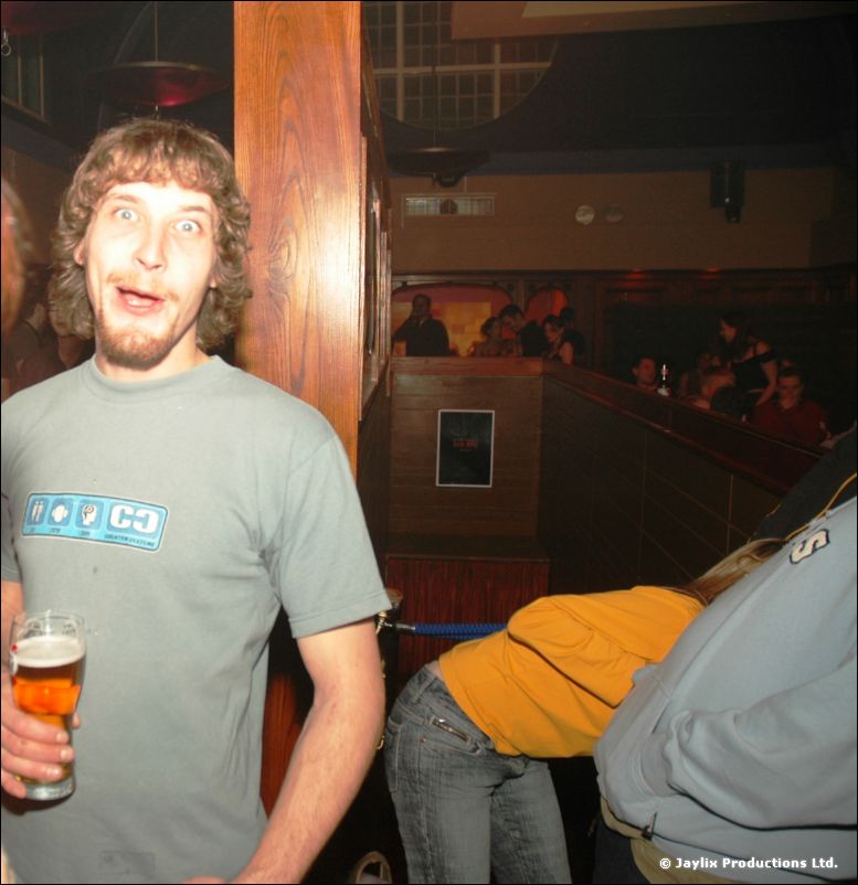 A man with curly hair in a grey t-shirt holds a pint and looks at the camera with wide eyes