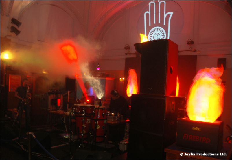 A dim hall with speakers, a glowing flame light and a projected white hand logo on the ceiling