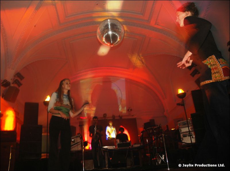 A vaulted hall with a mirror ball, a woman standing on a stage and a performer leaping at the edge