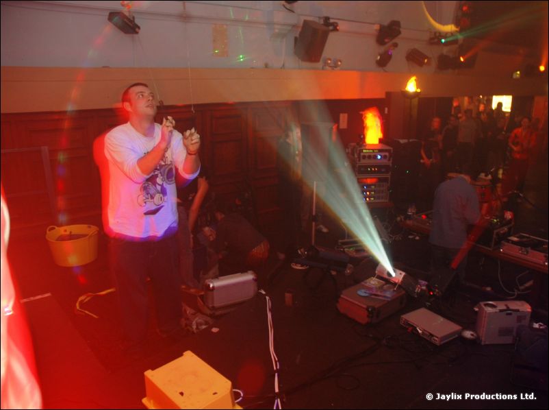 A person in a white t-shirt singing or performing on a stage, with light beams and equipment around