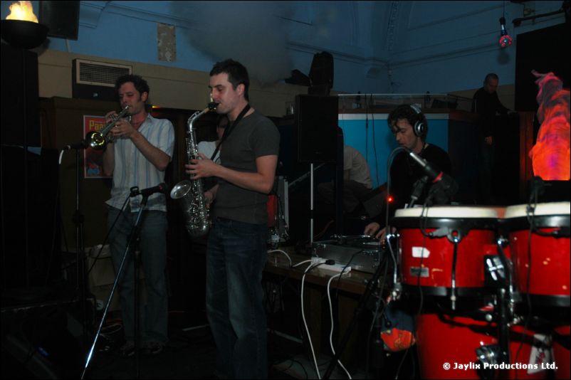 A trumpeter and a saxophonist stand by red conga drums with a person at the decks behind