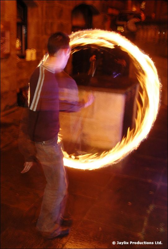 A person in a striped jacket spins a large ring of fire outside a stone building at night
