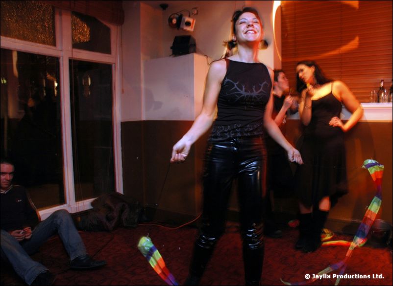A smiling woman in black leather trousers stands in a bar, rainbow ribbons on the floor around her