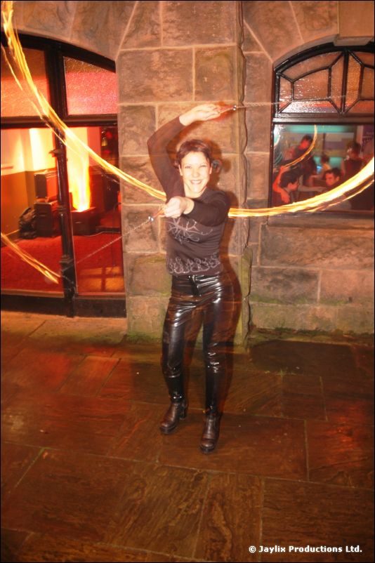 A smiling person in black leather trousers and boots spins fire trails outside a stone building
