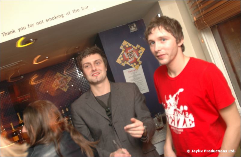 A man in a red T-shirt and a man in a grey jacket pose in a bar beside a no smoking sign