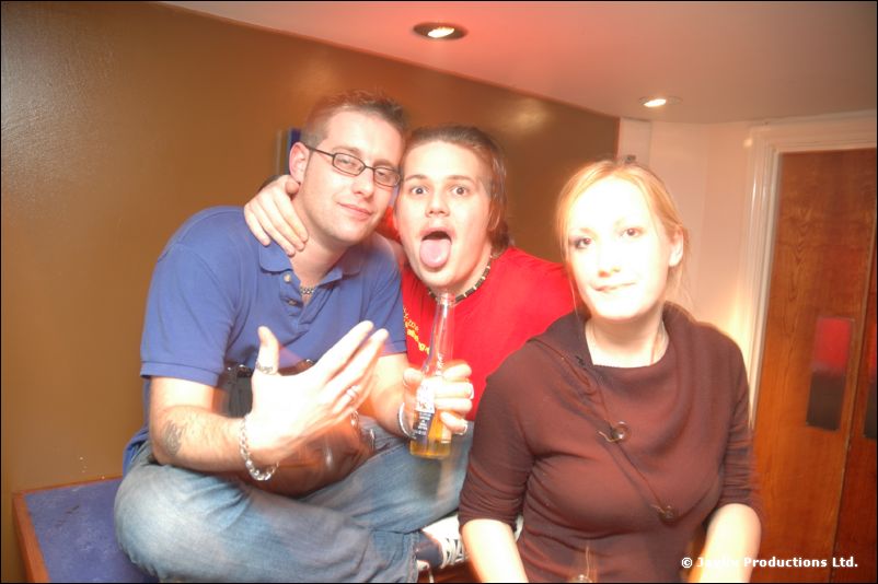 Two men and a woman sit close together, one man sticking out his tongue and holding a bottle