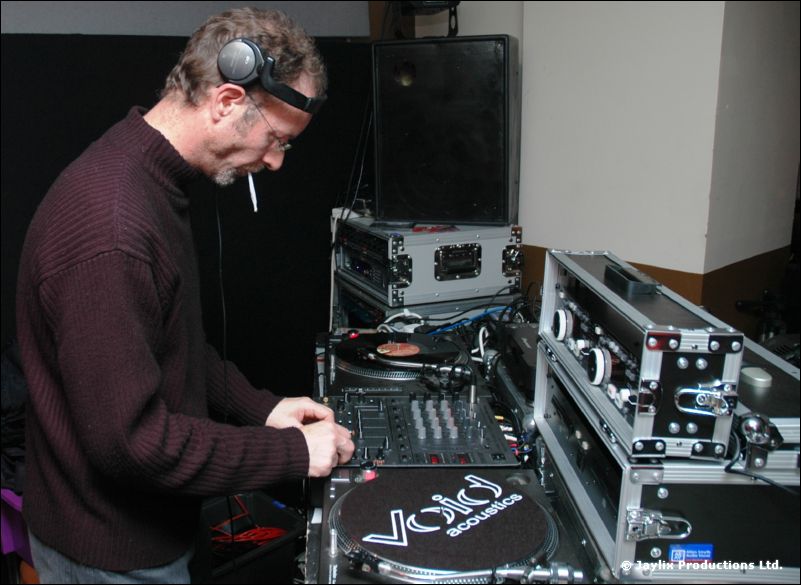 A DJ in a maroon jumper and headphones mixes at turntables with a cigarette in his mouth