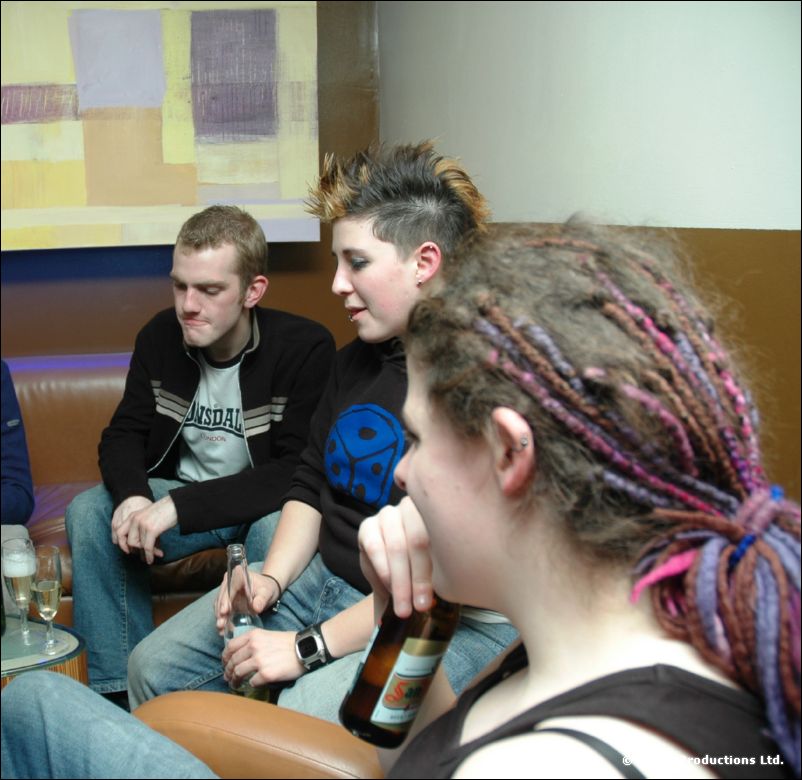 Two men and a person with purple dreadlocks sit on leather sofas with a beer bottle