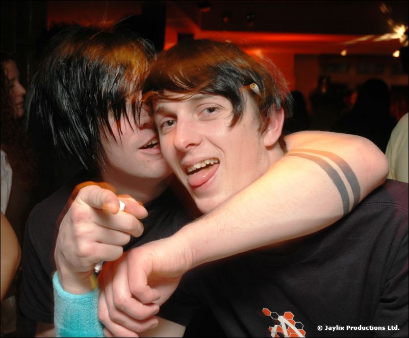 Two young people with dark hair hug and grin at the camera, one pointing