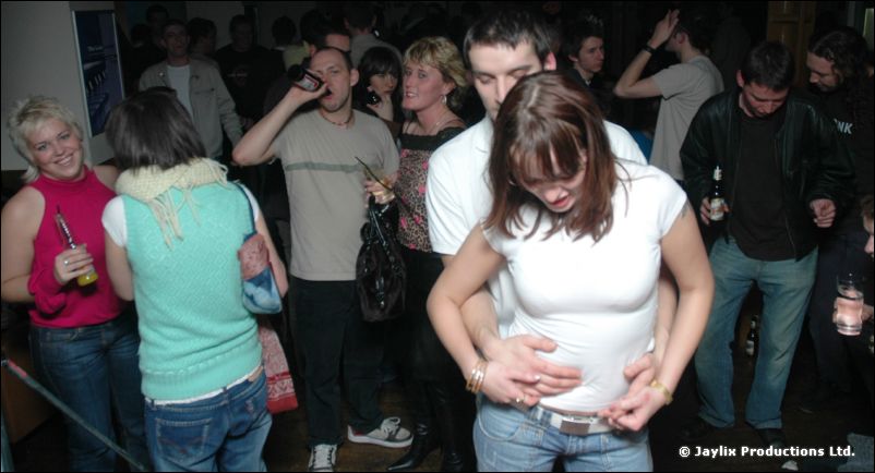 People dance and talk in a busy bar, one woman in white being held round the waist