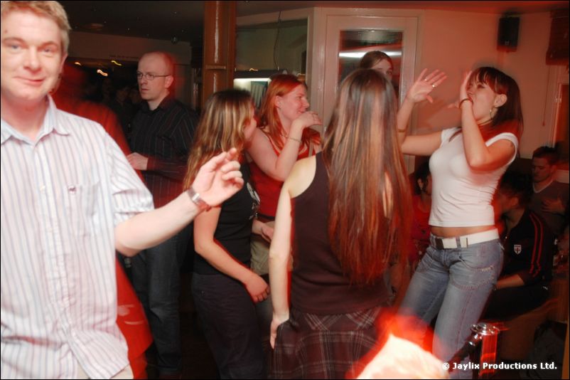 A man in a striped shirt dances among other dancers in a red-lit bar