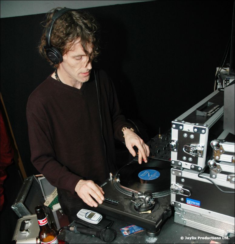 A DJ with curly hair and headphones mixes records at a turntable