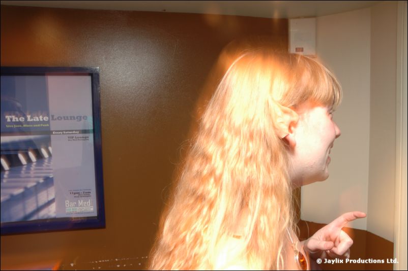 A woman with long blonde hair in profile points, beside a poster reading The Late Lounge