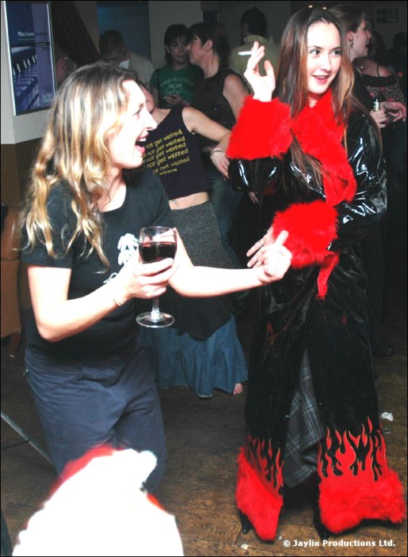 A woman with a glass of red wine laughs beside a woman in a black and red coat smoking
