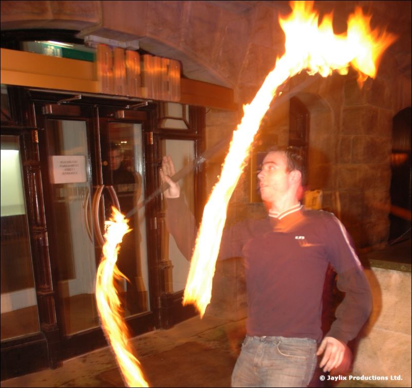A man in a dark top swings a burning fire staff outside the doors of a stone building at night