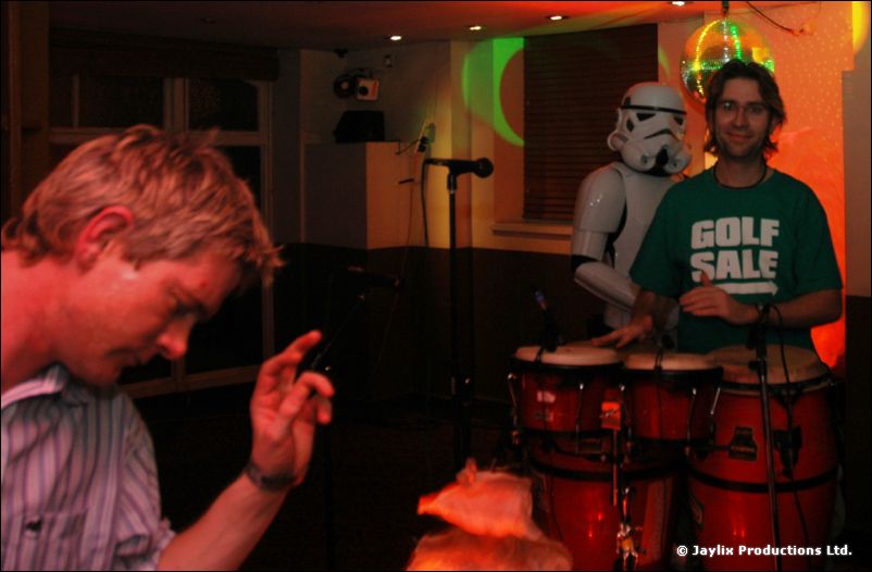 A man in a green Golf Sale T-shirt stands at conga drums beside a stormtrooper and a man in the foreground