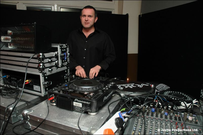 A DJ in black stands behind Pioneer decks and a mixer