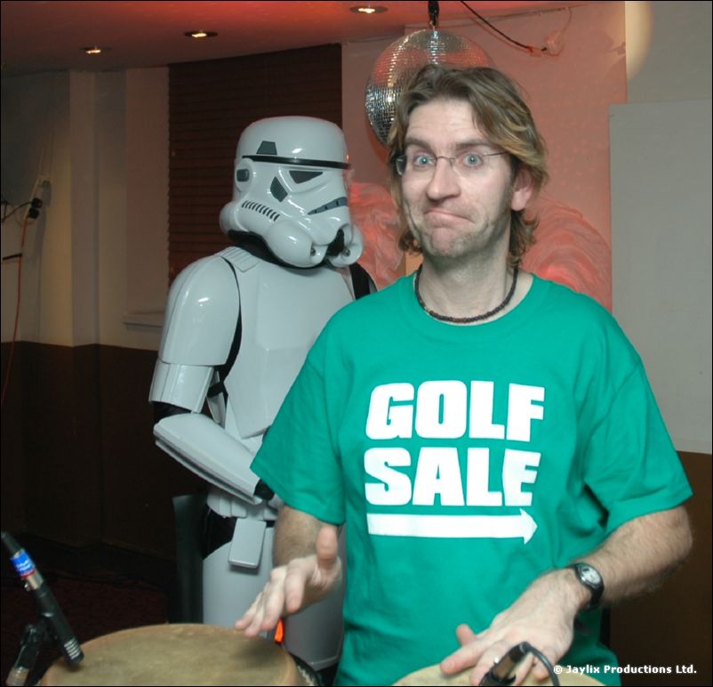 A man in a green Golf Sale T-shirt plays drums with a stormtrooper behind him