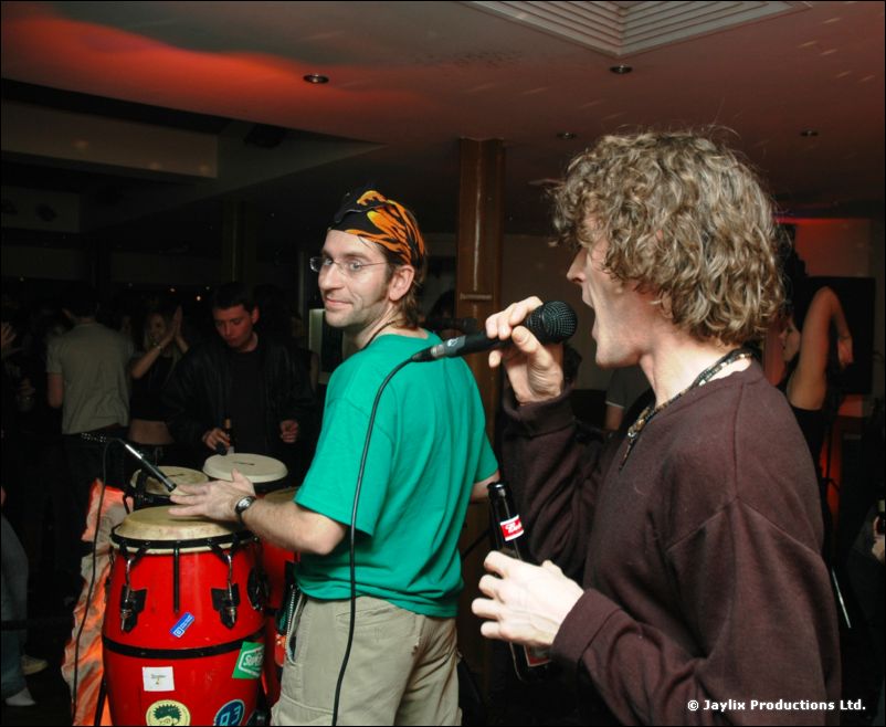 A man in a green top plays congas while another man with curly hair sings into a microphone