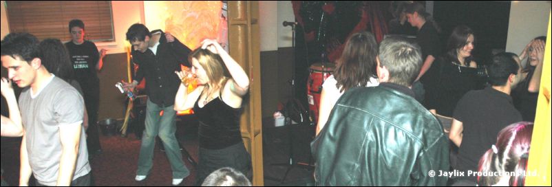 People dance and stand in a dimly lit bar, a man in a leather jacket in the foreground