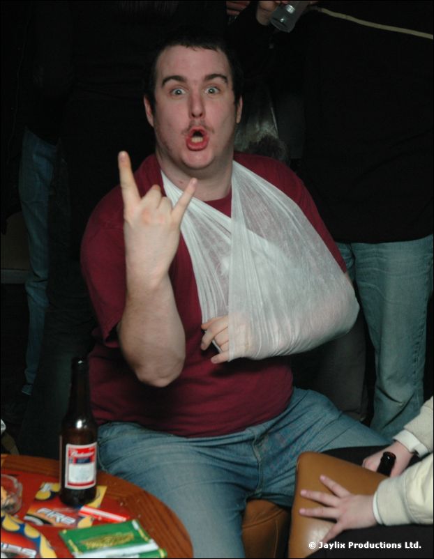 A man with his arm in a sling makes the rock sign and pulls a face beside a table with a beer bottle