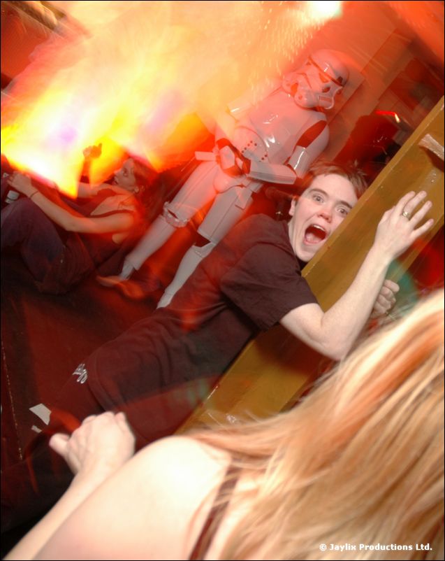 A person in a dark T-shirt shouts while leaning on a rail, with a stormtrooper and orange glow behind