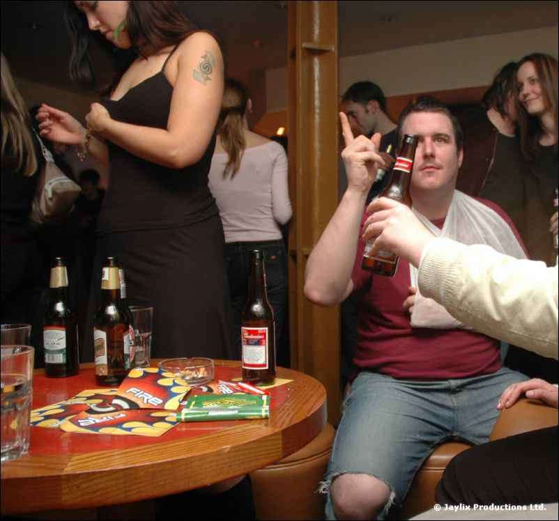 A man with an arm sling holds a beer bottle and makes a hand sign at a table with bottles