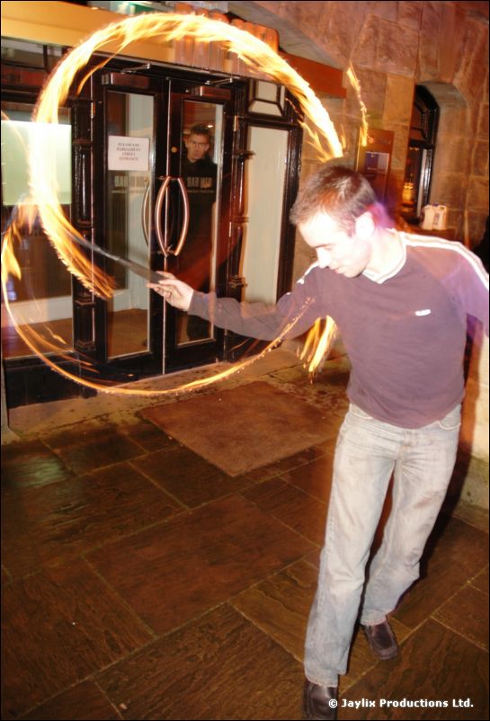 A man in a maroon top swings a ring of fire in front of glass doors at night