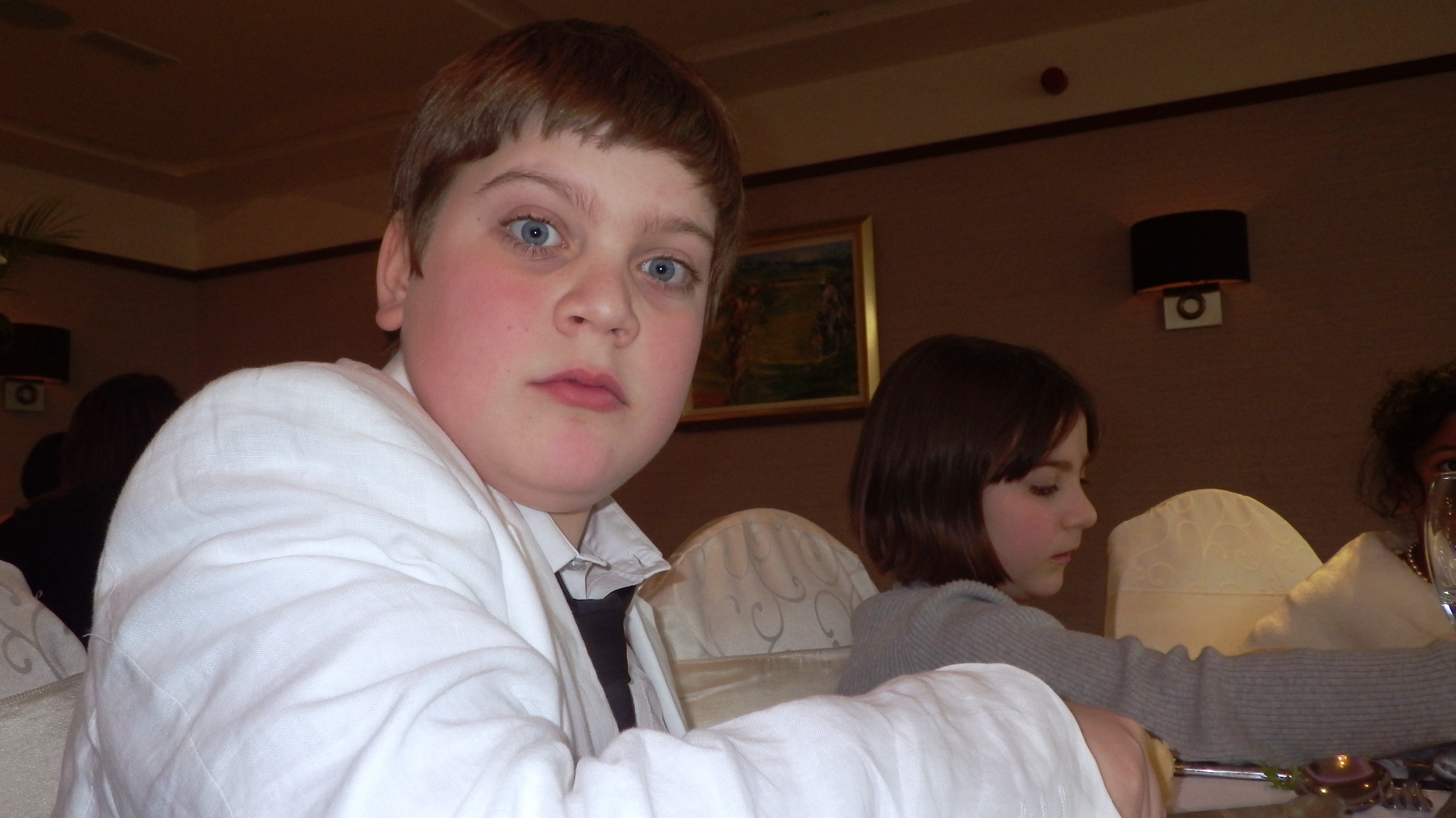 A child in a white jacket looks at the camera at a dining table, with another child behind