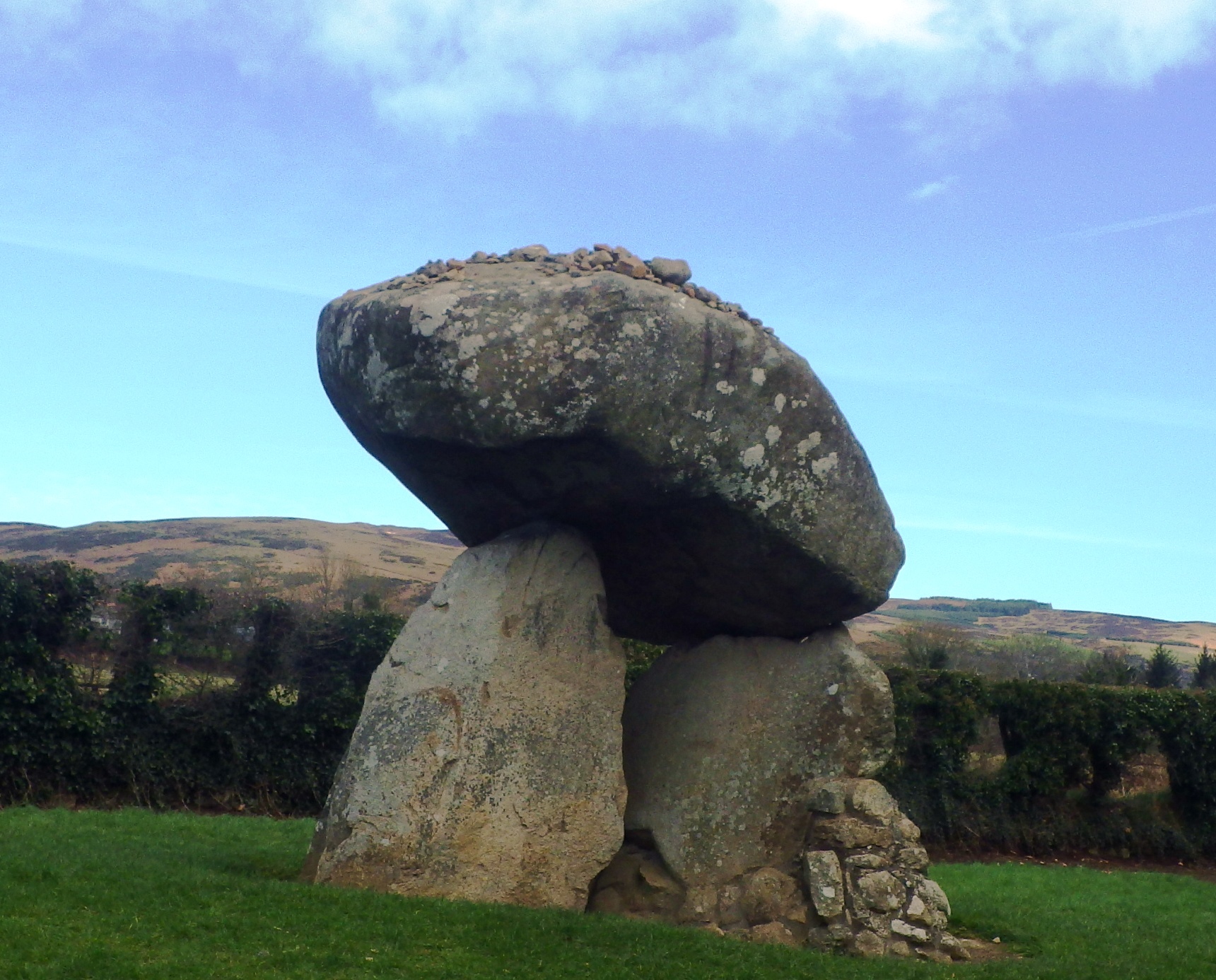 A large dolmen with a huge capstone on upright stones in a green field under a blue sky