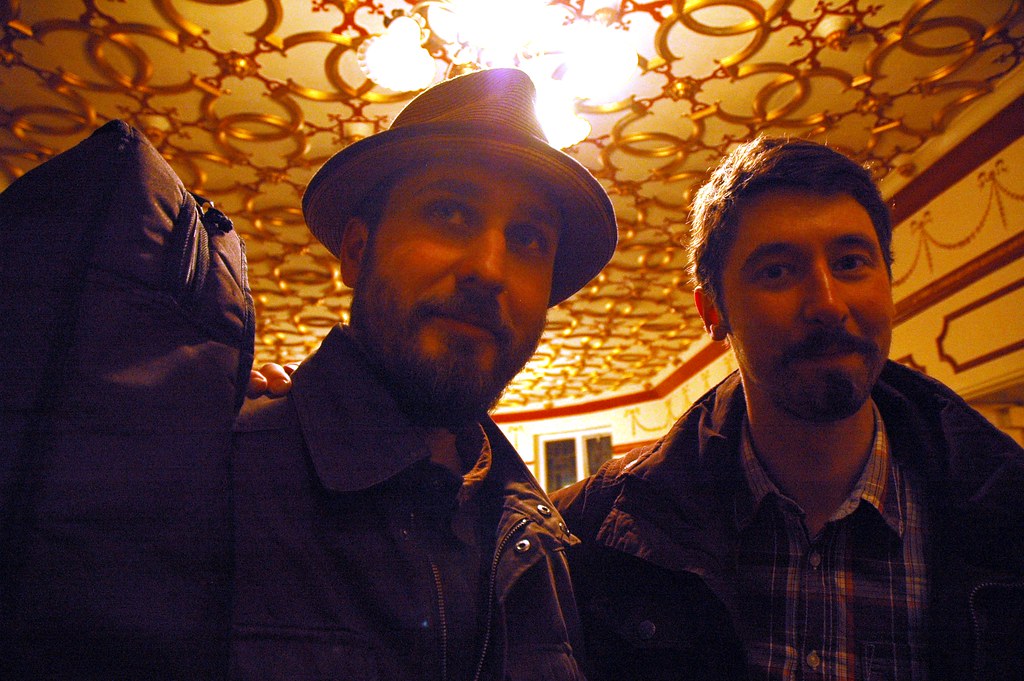 Two bearded men, one in a brimmed hat, pose close to the camera under an ornate gold ceiling