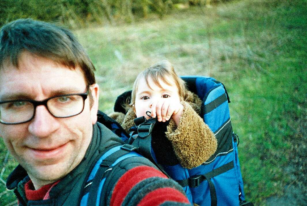 A man with glasses smiles in a selfie, with a toddler in a blue backpack carrier behind him
