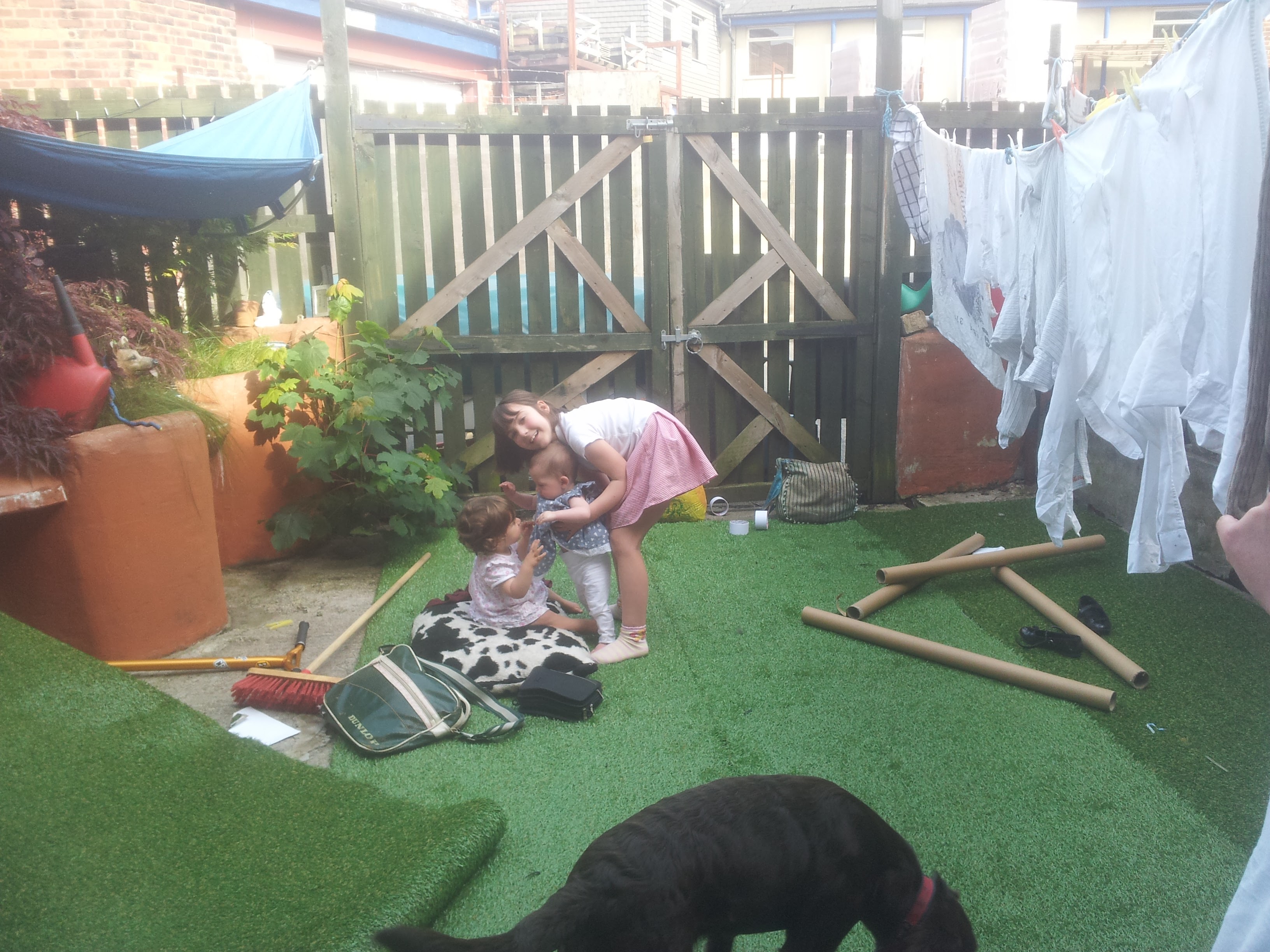 Children play on artificial turf in a small back yard, with washing on a line and a black dog