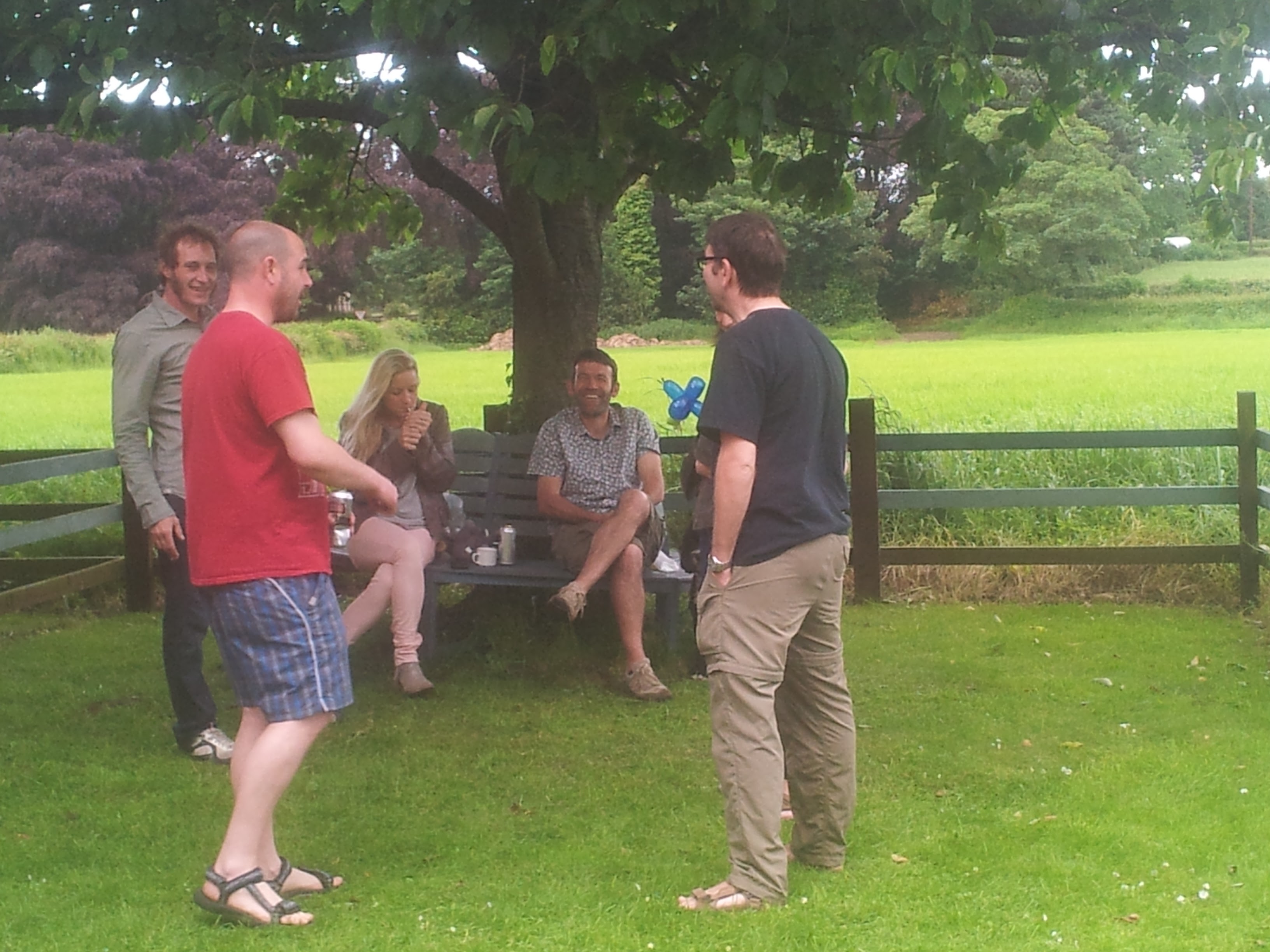 Five adults chat on a lawn under a tree beside a wooden fence
